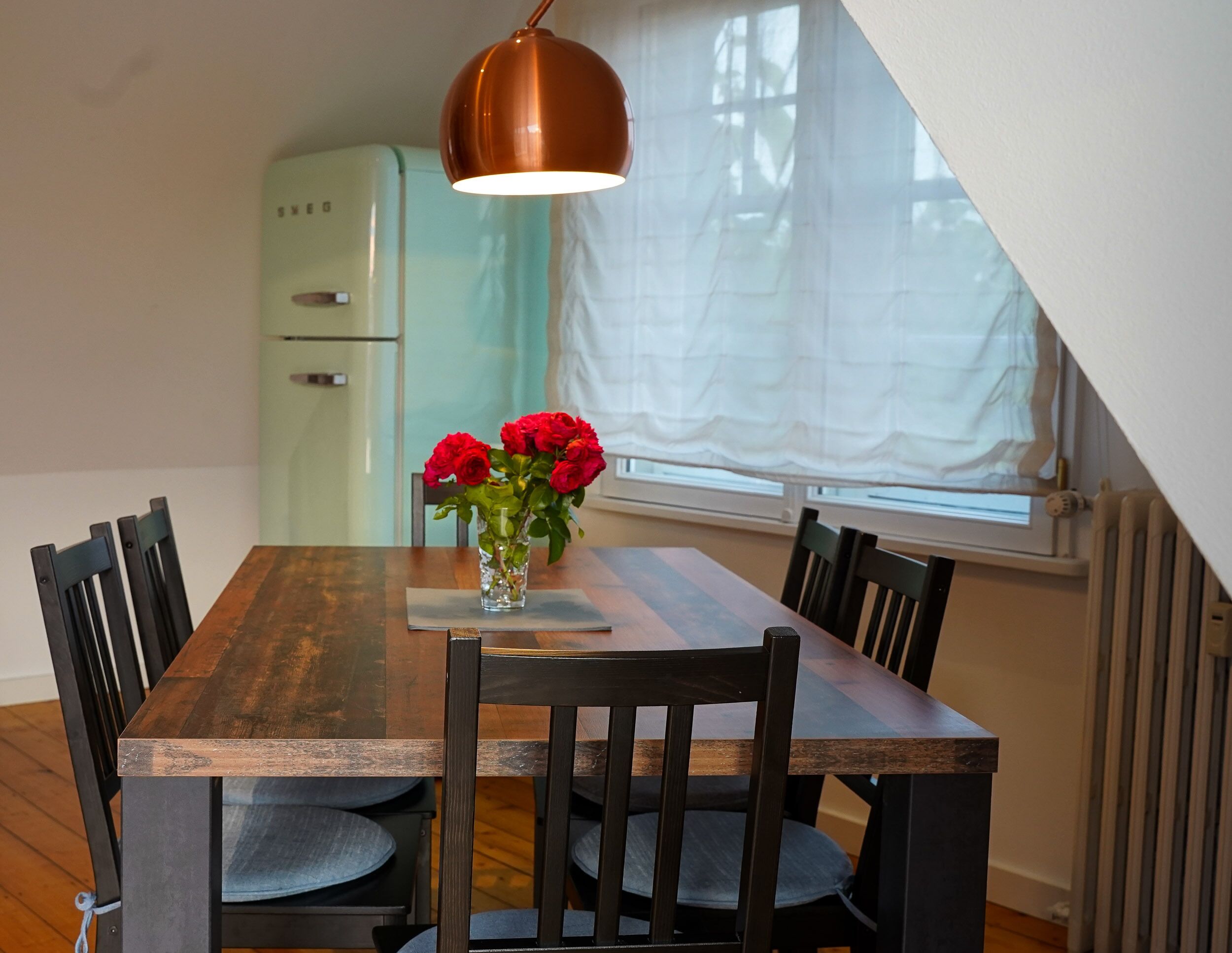 A dining area with a wooden table, four chairs, a vase with red flowers, a copper pendant light, a vintage green refrigerator, and a window with white curtains.