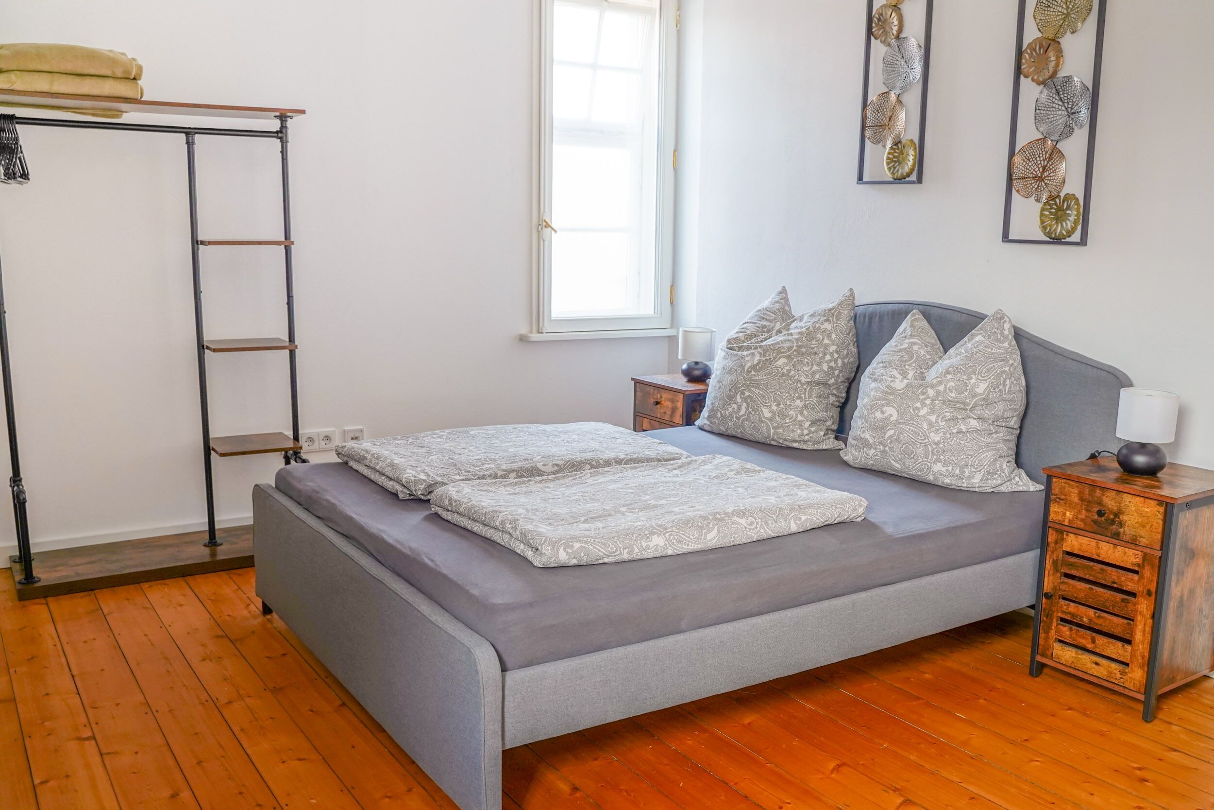 A bedroom with a grey bed, wooden floor, a metal ladder, a bedside table with a lamp, and decorative wall art.