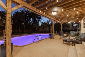 Ein Außenpoolbereich mit einer Holzpergola, Liegestühlen und einem Pool mit violetter Beleuchtung.
