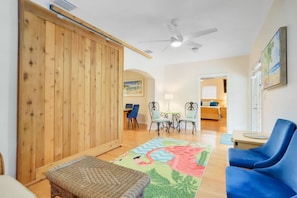 A room with a wooden partition, a ceiling fan, a dining area with chairs, a blue sofa, and a colorful rug.
