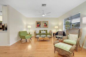 Una sala de estar con un sillón verde, un sofá, una silla de mimbre, una mesa de centro y dos lámparas de pared.
