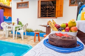 Un gâteau au chocolat sur une assiette bleue, posé sur une nappe blanche à motifs de dentelle, entouré de fruits et avec une piscine en arrière-plan.