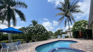 A swimming pool with a tiled edge, surrounded by a brick patio, palm trees, and a blue umbrella. - A Tropical 2 Bedroom/2 Bath House w/ Pool and Dock (Osprey)