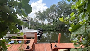 A wooden dock with a metal railing, a boat docked at the pier, and a calm water body with trees in the background. - A Tropical 2 Bedroom/2 Bath House w/ Pool and Dock (Osprey)