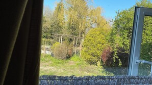 A view through a window with a curtain, featuring a garden with various trees and a wooden pergola. - 1 bedroom accommodation in Booton, Reepham (Booton, Reepham)