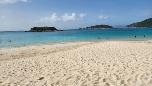 Une plage de sable fin, avec une eau d’un bleu limpide et des formations rocheuses visibles au loin.