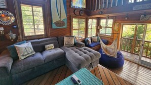 L'intérieur d'un chalet accueillant, avec un grand canapé d'angle gris, un hamac et une terrasse en bois offrant une vue sur la verdure.