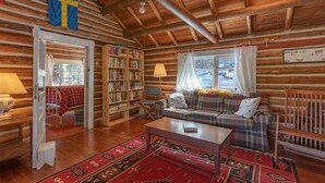 Ein Blockhaus-Interieur mit Holzbalkendecke, einem Karo-Sofa, einem Bücherregal und einem rot gemusterten Teppich.