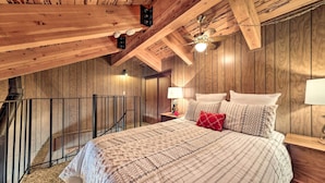 Um quarto aconchegante com teto de madeira, uma cama com roupa de cama estampada, uma mesa de cabeceira com abajur e uma escada.