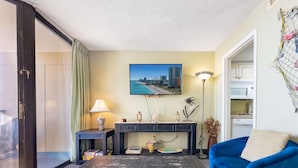 A living room with a blue sofa, a coffee table, a lamp, a picture of a beach, and a kitchenette. - Sand Dunes 2626 (Myrtle Beach)