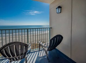 A balcony with a view of the beach and ocean, two chairs, and a wall-mounted light fixture.