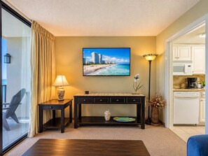 A hotel room with a large window, a flat-screen TV, a dining area with a table and chairs, and a kitchenette.