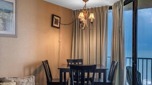 A dining area with a table and chairs, a chandelier, a picture on the wall, and a view through a window. - Oceanfront Retreat | Sand Dunes Twenty-Six Twenty-Six (Myrtle Beach)