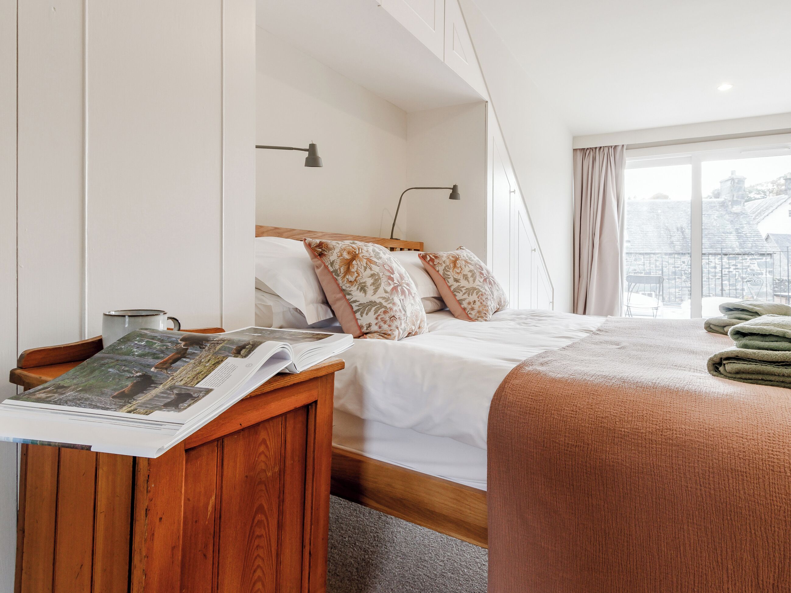 A hotel room with a bed, a bedside table with a magazine, a window with curtains, and a view of the city.