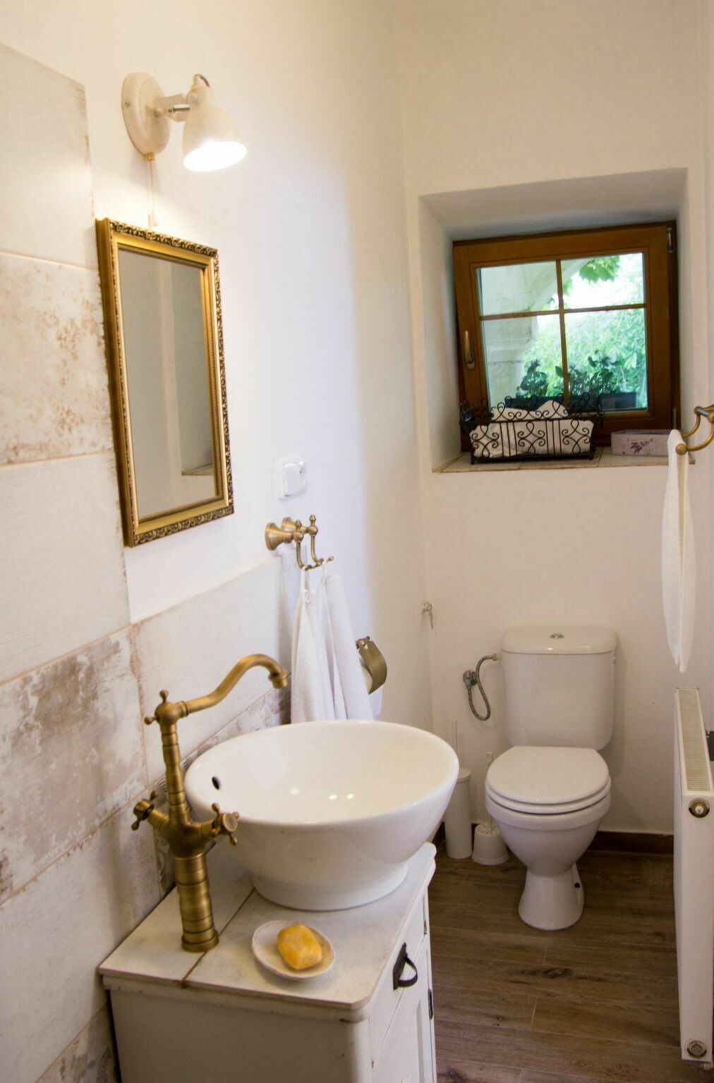 A bathroom with a white sink, gold faucet, and a gold-framed mirror. There is a window above the toilet.