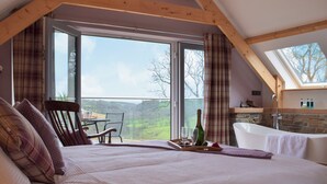 A bedroom with a large bed, a dining table with chairs, a view through a glass door, and a stone wall with a bathtub. - 4 bedroom accommodation in Llanboidy, near Laugharne (Llanboidy, near Laugharne)