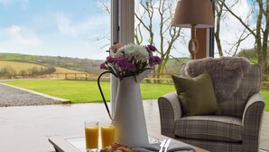 A breakfast setup with croissants, juice, and a vase of flowers on a table by a window overlooking a rural landscape. - 4 bedroom accommodation in Llanboidy, near Laugharne (Llanboidy, near Laugharne)