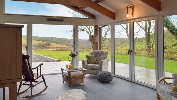 A spacious room with a large glass door leading to a scenic outdoor view, a wooden rocking chair, a wicker chair, and a small table with a vase of flowers. - 4 bedroom accommodation in Llanboidy, near Laugharne (Llanboidy, near Laugharne)