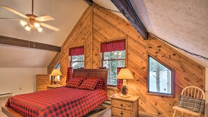 Een slaapkamer met een schuine kap, houten wanden, een bed met een rode geruite plaid, twee nachtkastjes met lampen, en een stoel met een ruitkussen.