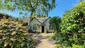 A quaint cottage with a thatched roof, surrounded by lush greenery and a gravel path leading to the entrance.