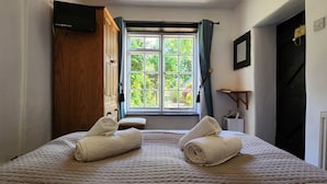 A bedroom with a bed, a window with curtains, a wooden wardrobe, and a television mounted on the wall.