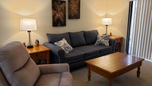 Ein Wohnzimmer mit einer Couch, einem Couchtisch und zwei Lampen. An den Wänden hängen zwei Gemälde und ein Fenster mit Jalousien.