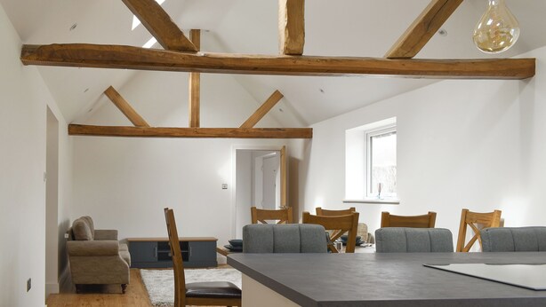 Un loft espacioso con vigas de madera a la vista, una zona de comedor con sofás grises y un ventanal que proporciona luz natural.