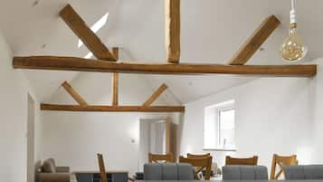 A spacious loft with exposed wooden beams, a dining area with grey sofas, and a large window providing natural light.