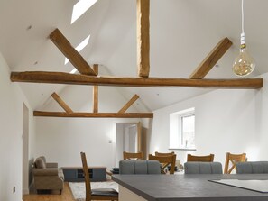 Ein geräumiger Loft mit freiliegenden Holzbalken, einer Essecke mit grauen Sofas und einem großen Fenster, das natürliches Licht hereinlässt.