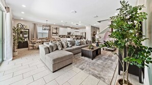 En moderne stue med en grå hjørnesofa, et sofabord og et spiseområde med et træbord og stole. Der er et køkken i baggrunden med hvide skabe og en lysekrone.
