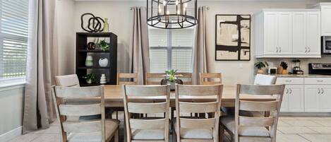 Een eethoek met een houten tafel en stoelen, een hanglamp, een boekenplank en een keuken op de achtergrond.