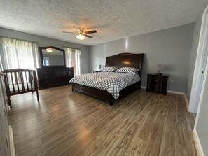 Une chambre avec un plancher en bois, un lit recouvert d’une couvre-lit à motifs, un ventilateur de plafond et une commode.