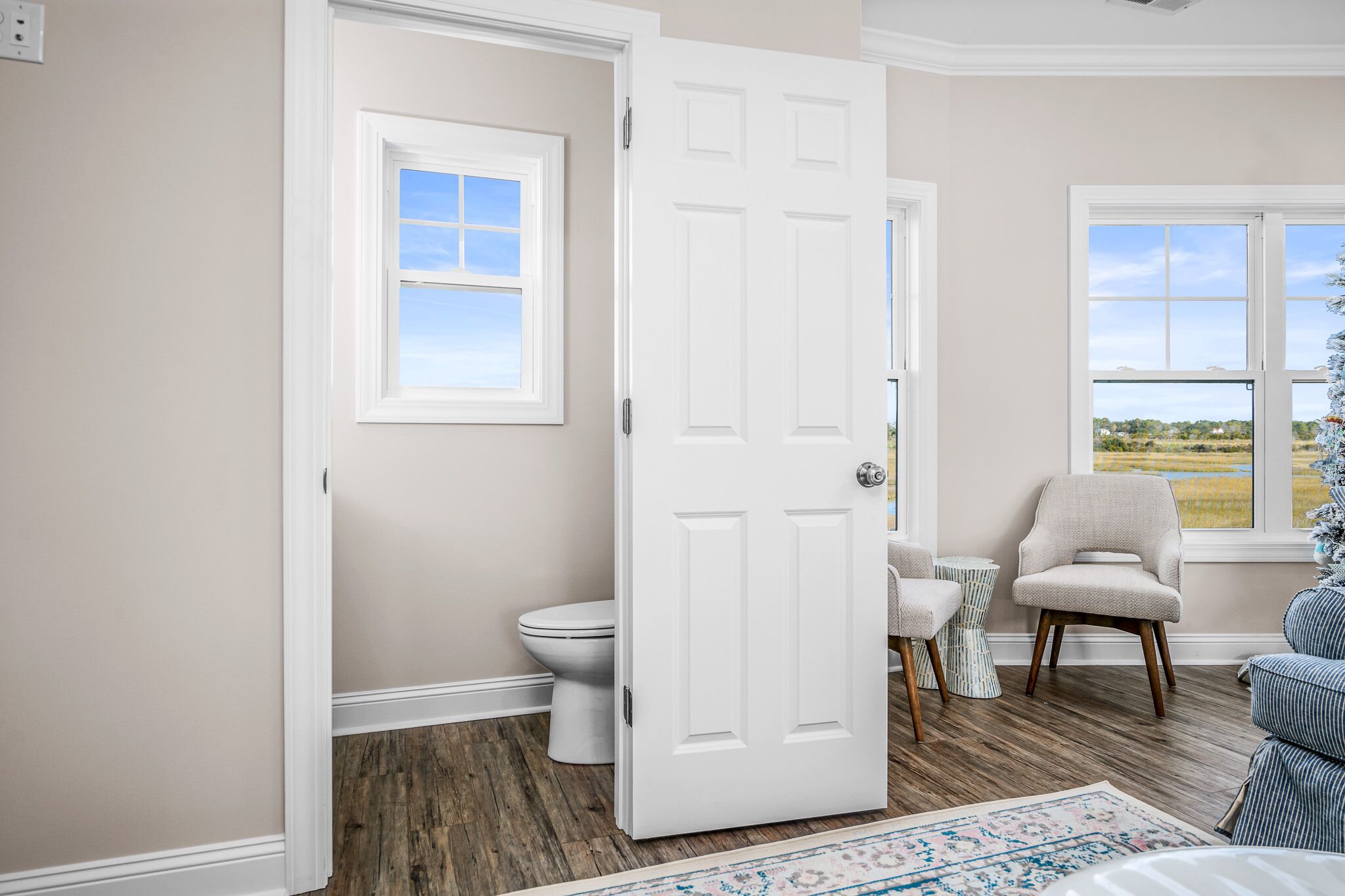 A bathroom with a toilet, a white door, a window, and a chair.