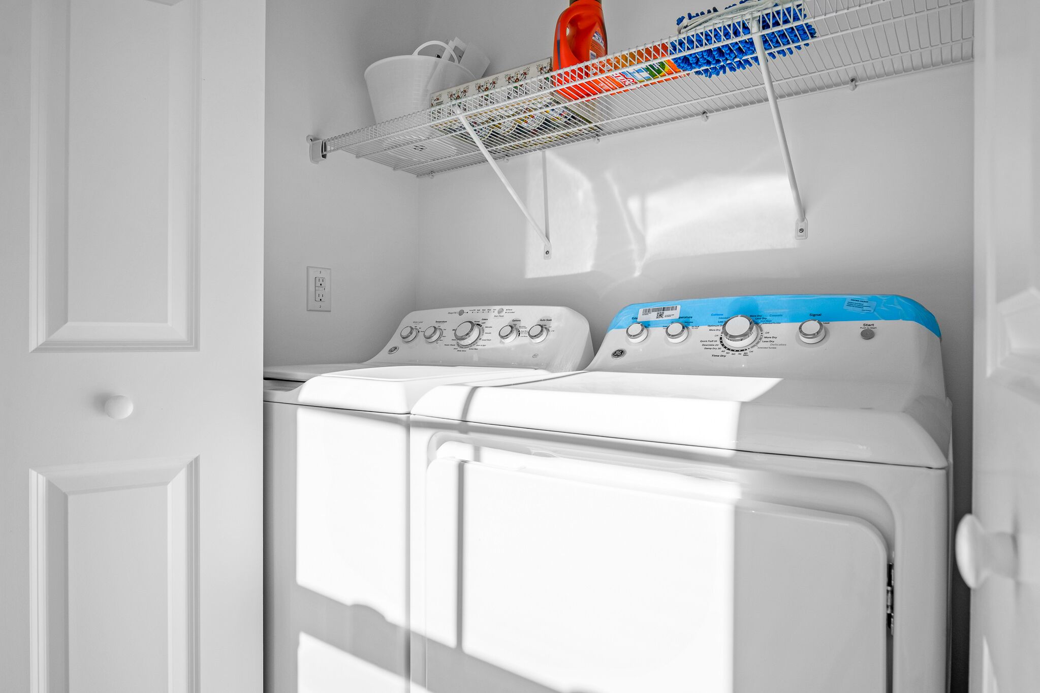 A front-loading washer and dryer in a laundry room with a shelf above containing cleaning supplies.