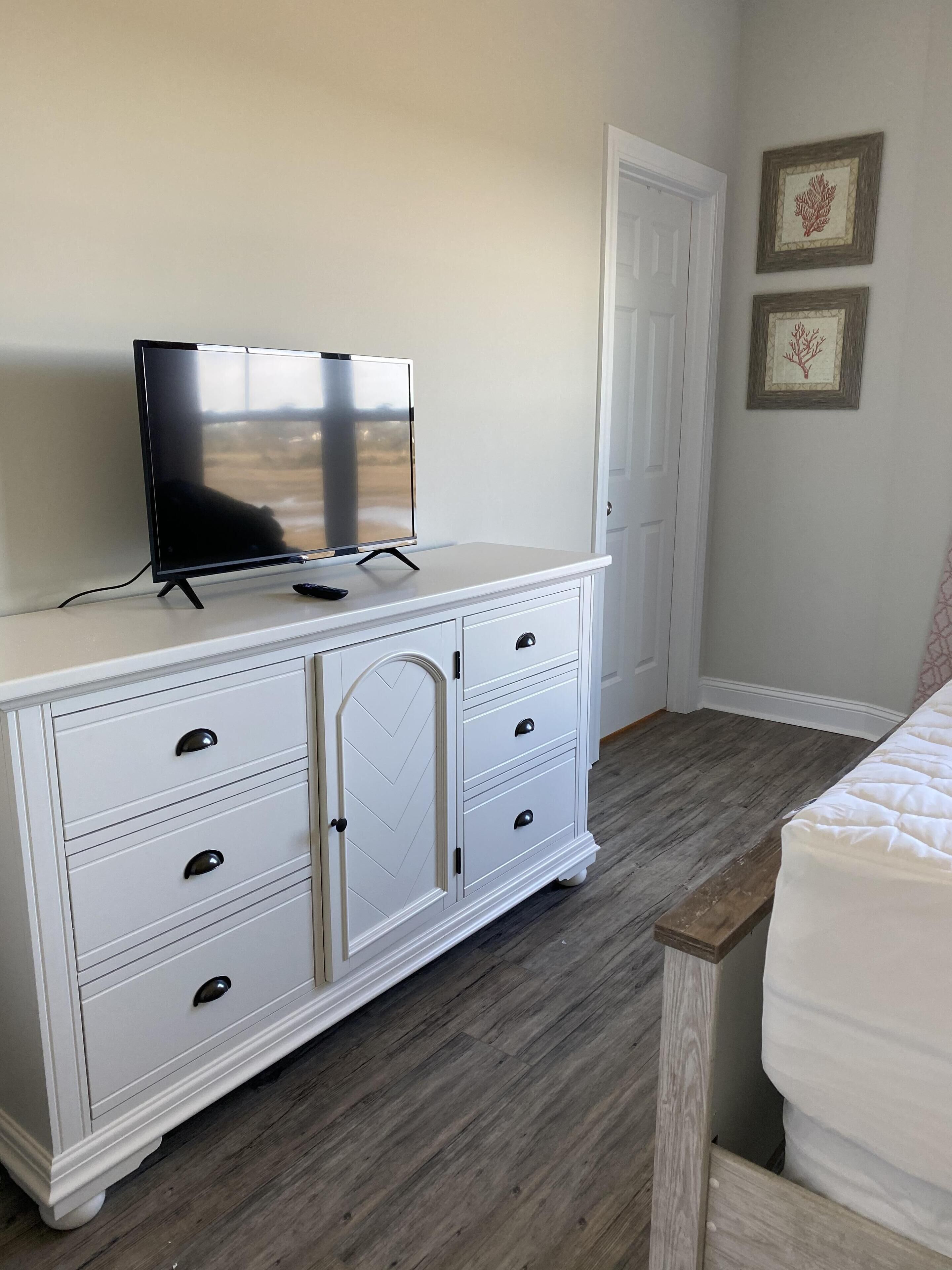 Une chambre avec une commode blanche, un téléviseur posé dessus, un banc en bois et une porte.