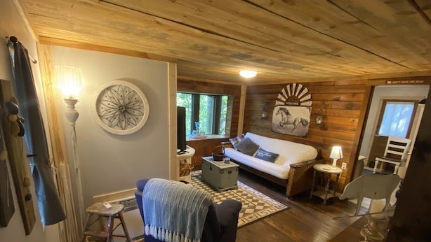 A cozy cabin interior with a wooden ceiling, a white sofa with a blue throw, a small table with a lamp, and a decorative wall piece.