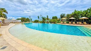 Una gran piscina curvada con sillas de descanso y sombrillas, rodeada de palmeras y follaje.