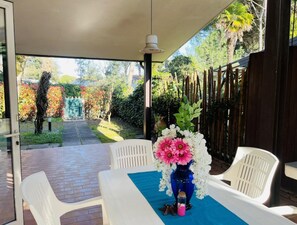 A covered patio with a table set for two, a vase of flowers, and a view of a garden. - Lovely Villa With Spacious Garden (San Michele al Tagliamento)