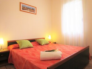 Une chambre d’hôtel avec un lit, des oreillers, une table de chevet, une lampe, une fenêtre avec des rideaux et un tableau encadré au mur.