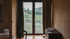 Ein Zimmer mit Balkon, einem Sessel, einem kleinen Tisch mit einem Glasbehälter und einem fenster mit Vorhängen.