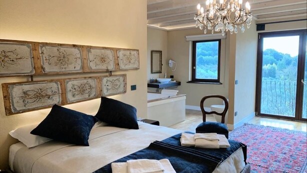 Ein Schlafzimmer mit Bett, einer Kristallleuchter, Kunstwerken an der Wand, einem Fenster und Blick ins Freie.
