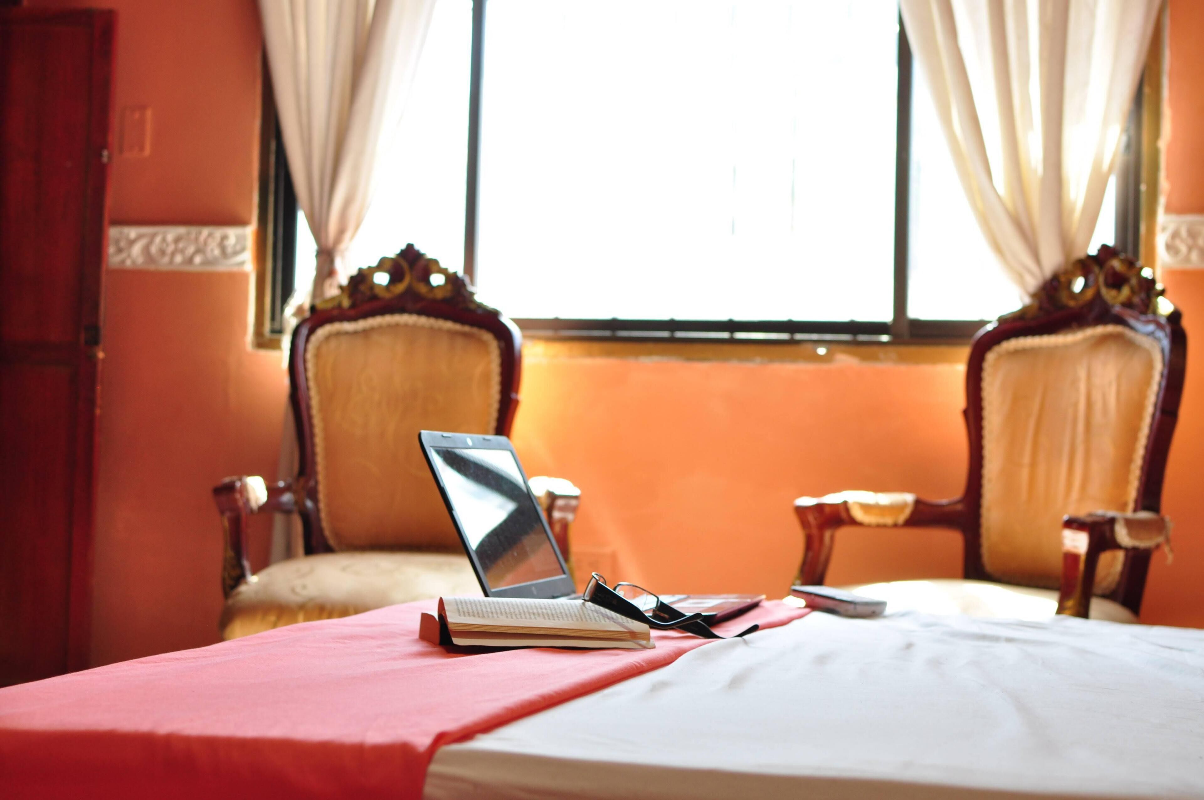 Una habitación con una cama, una computadora portátil y un libro sobre una mesa. Hay dos sillas y una ventana con cortinas.