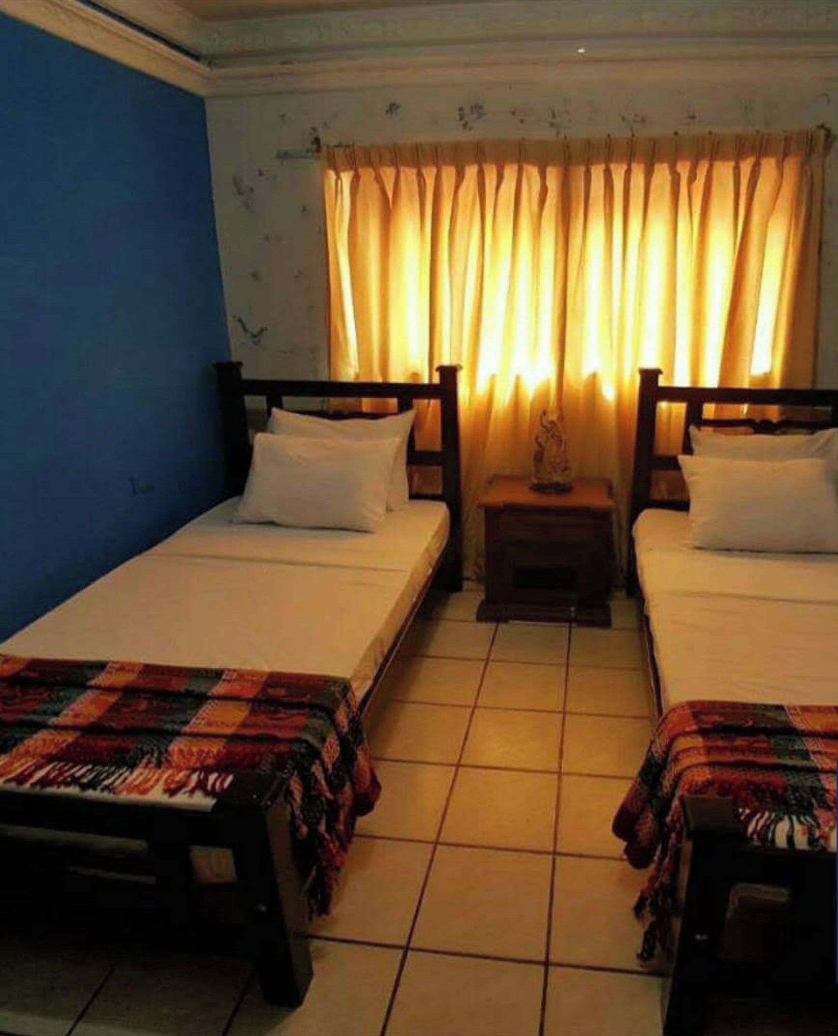 A room with two single beds, a small bedside table with a lamp, and a window with curtains.