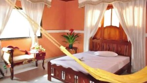 A bedroom with a wooden bed, white curtains, a nightstand, and a potted plant. - Hostal Hogar Buenaventura (Santa Marta)