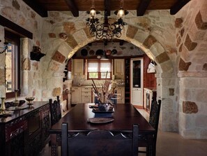 A stone-walled dining area with a wooden table and chairs, a chandelier, and a view into the kitchen.