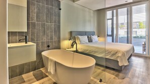 A modern bedroom with a large bed, a freestanding bathtub, and a bathroom with a mirror and sink. - Luxury 2 Bedroom With Balcony (Cape Town)