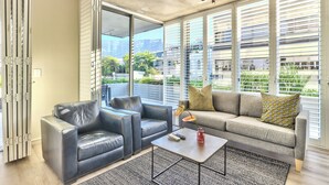 A modern living room with a sofa, two armchairs, and a coffee table. Large windows offer views of buildings and greenery. - Luxury 2 Bedroom With Balcony (Cape Town)