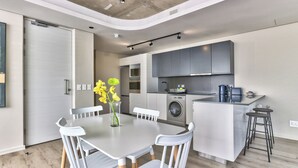 A modern kitchen with grey cabinets, a built-in oven, and a washing machine. There is a dining area with a white table and chairs, and a vase with yellow flowers. - Luxury 2 Bedroom With Balcony (Cape Town)