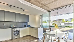 A modern kitchen with a built-in washer, large windows, and a dining area with white chairs and a table. - Luxury 2 Bedroom With Balcony (Cape Town)