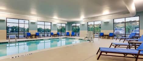 A swimming pool with blue lounge chairs and a view of the outdoors through large windows.